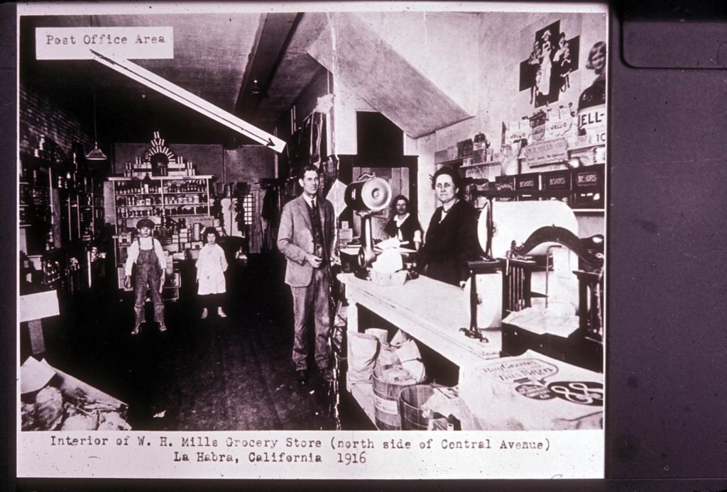 Interior of the Mills grocery store, located on the north side of Central Avenue, in the Wester Building, La Habra, Calif., in 1916. Between 1916 and 1920 the store included a post office area. At the counter are Mr. and Mrs. Mills and assistant, Katie Terwiliger. Although the photograph label identifies this as the W.H. Mills store, other sources identify it as the store operated by W.H.'s son, W.P. (Phillip) Mills.