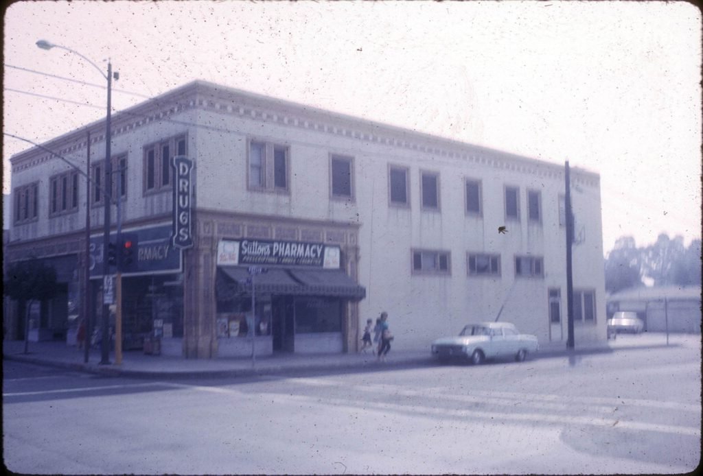 Image of the Sutton's Pharmacy (exterior) (corner of present La Habra Blvd.), with people walking to a car.